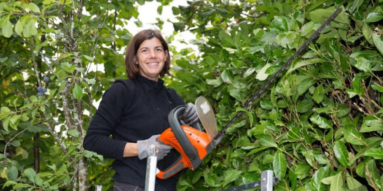 Smiling woman holding an electric hedge trimmer in front of a hedge